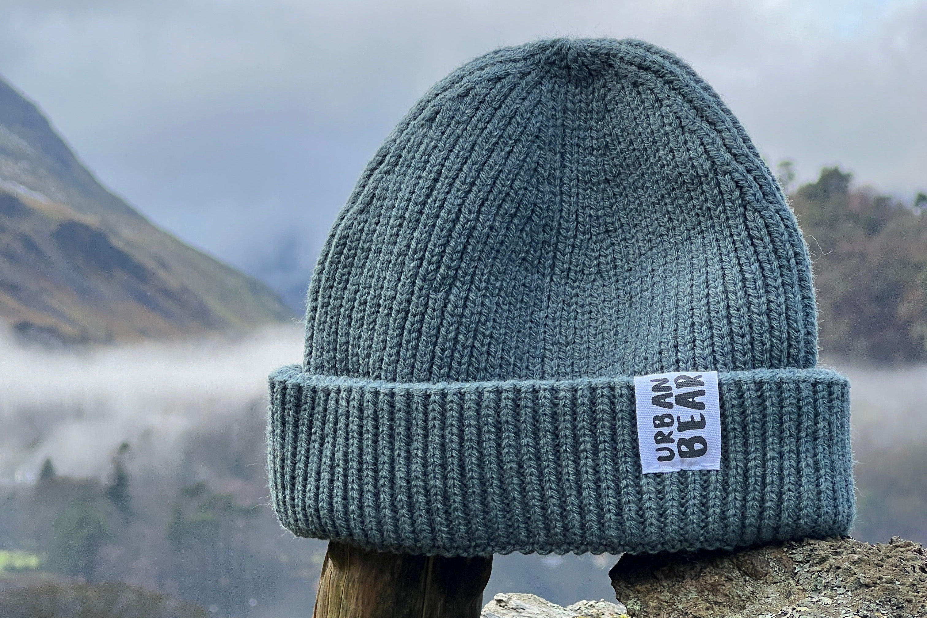 A gray ribbed knit beanie hat with a cuffed design, displayed outdoors with a natural background.