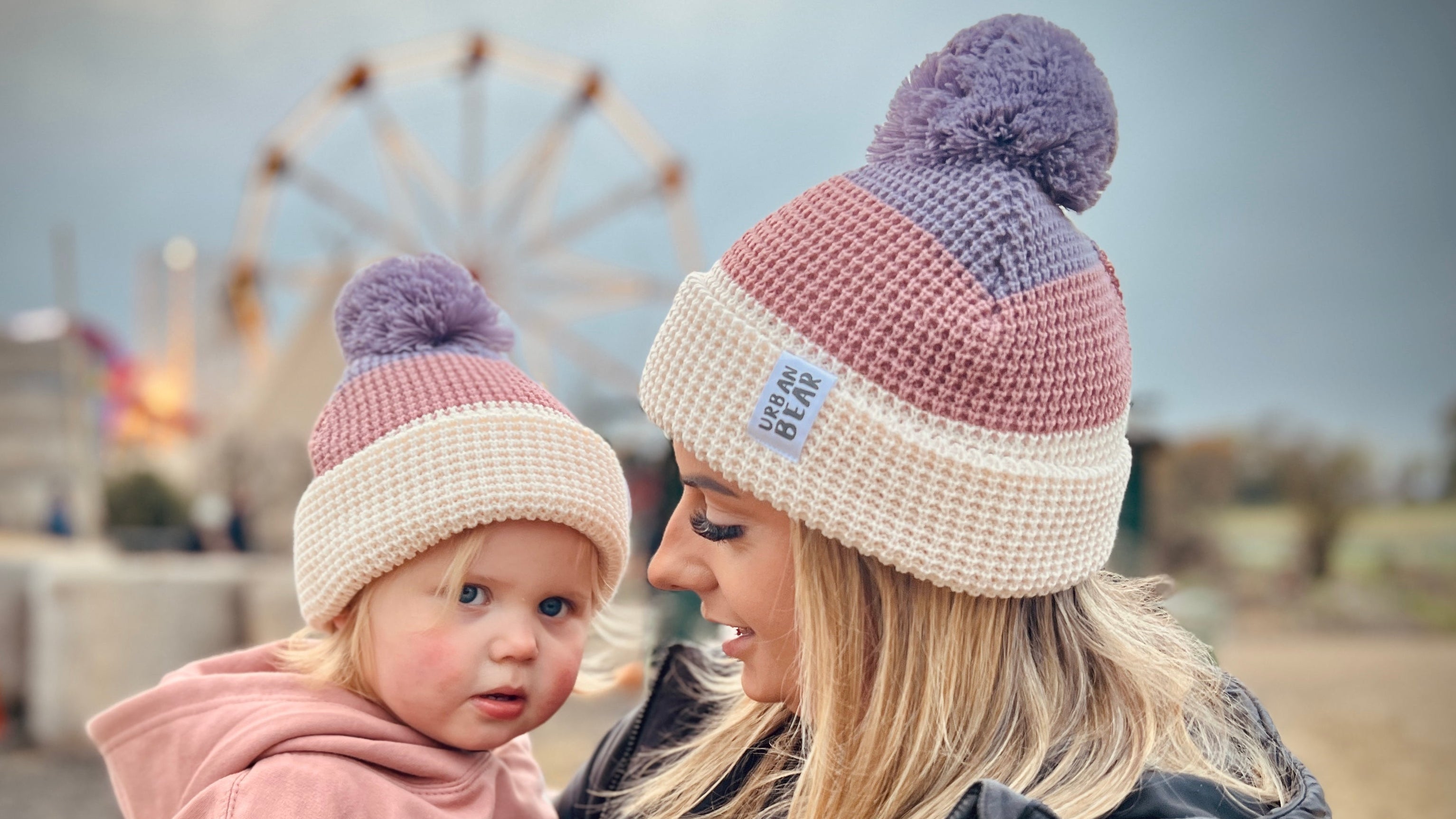 An adult and a child wearing matching pink and purple waffle knit hats with pom poms, standing outdoors with a carnival in the background.