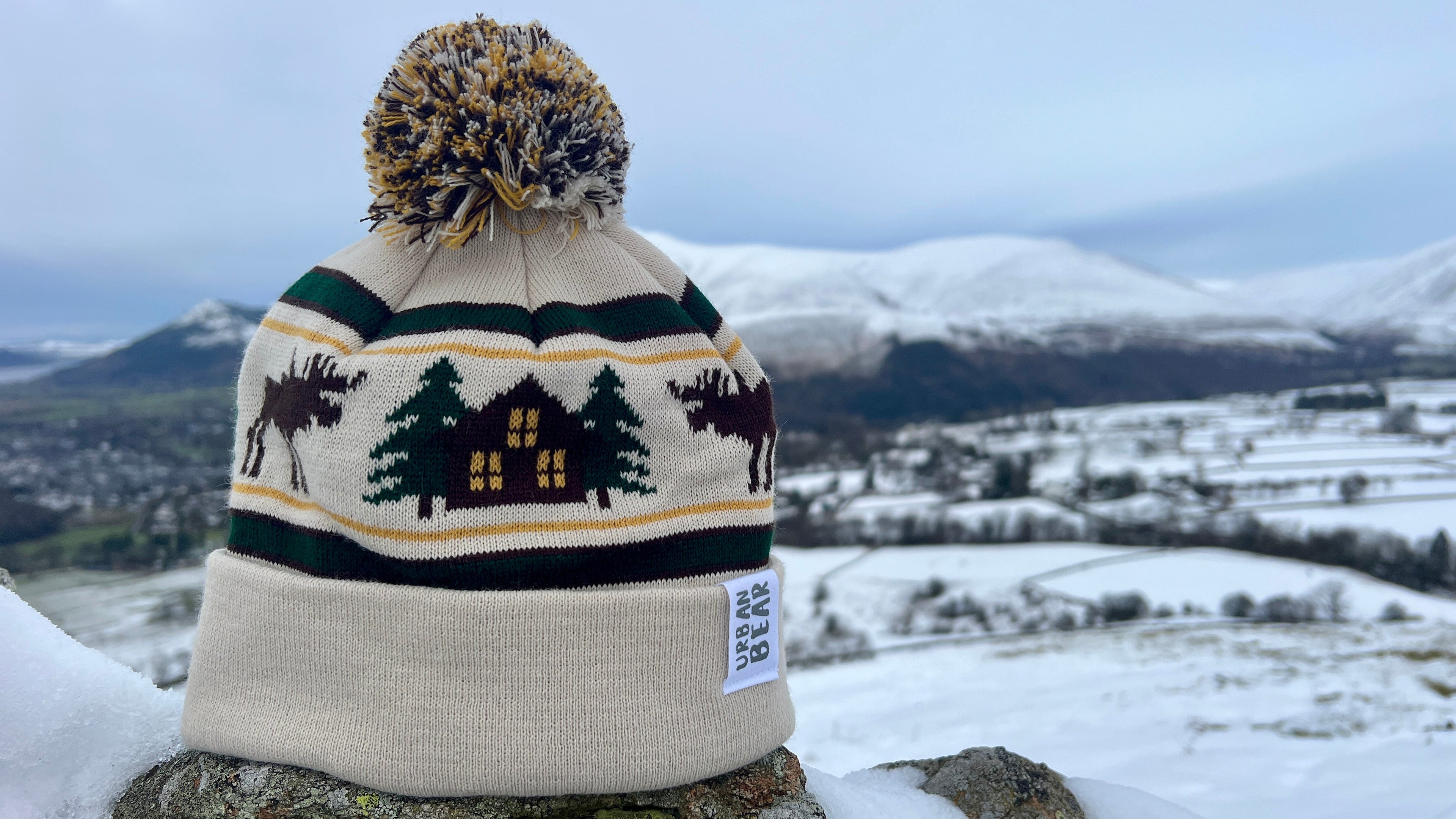 Cream bobble hat with log cabin and reindeers with snowy mountain background