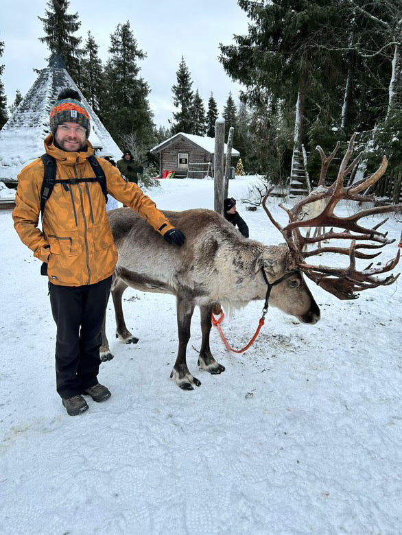A man in a yellow-orange winter jacket and black pants stands in a snowy outdoor setting next to a large reindeer with large antlers. Trees, cabins, and snow are visible in the background.
