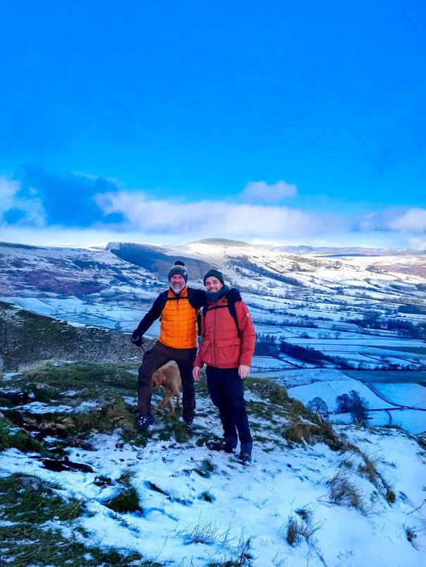Two people standing on a snowy hilltop with a dog, wearing winter jackets and hats, with a snow-covered valley and blue sky in the background.
