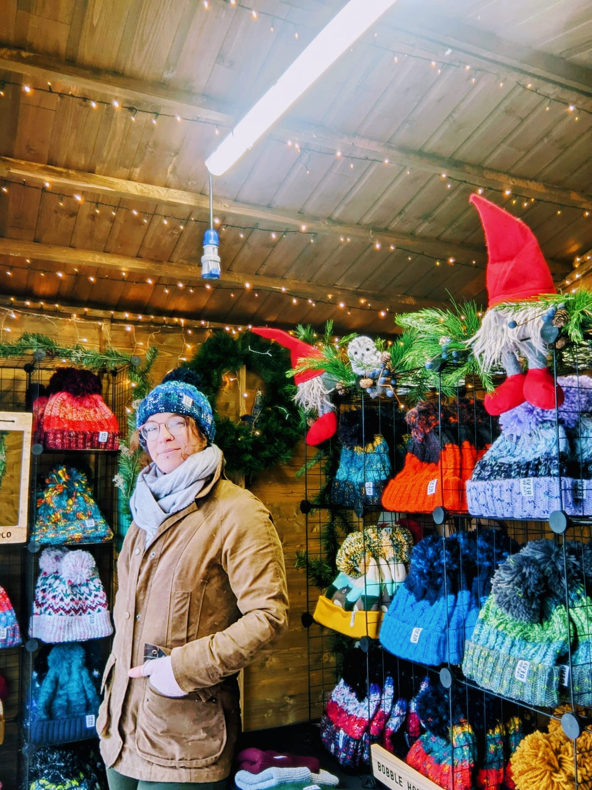 A woman wearing a blue knit beanie, gray scarf, and brown jacket stands inside a rustic shop filled with colorful knit hats, gloves, and gnome decorations hanging on wooden walls.