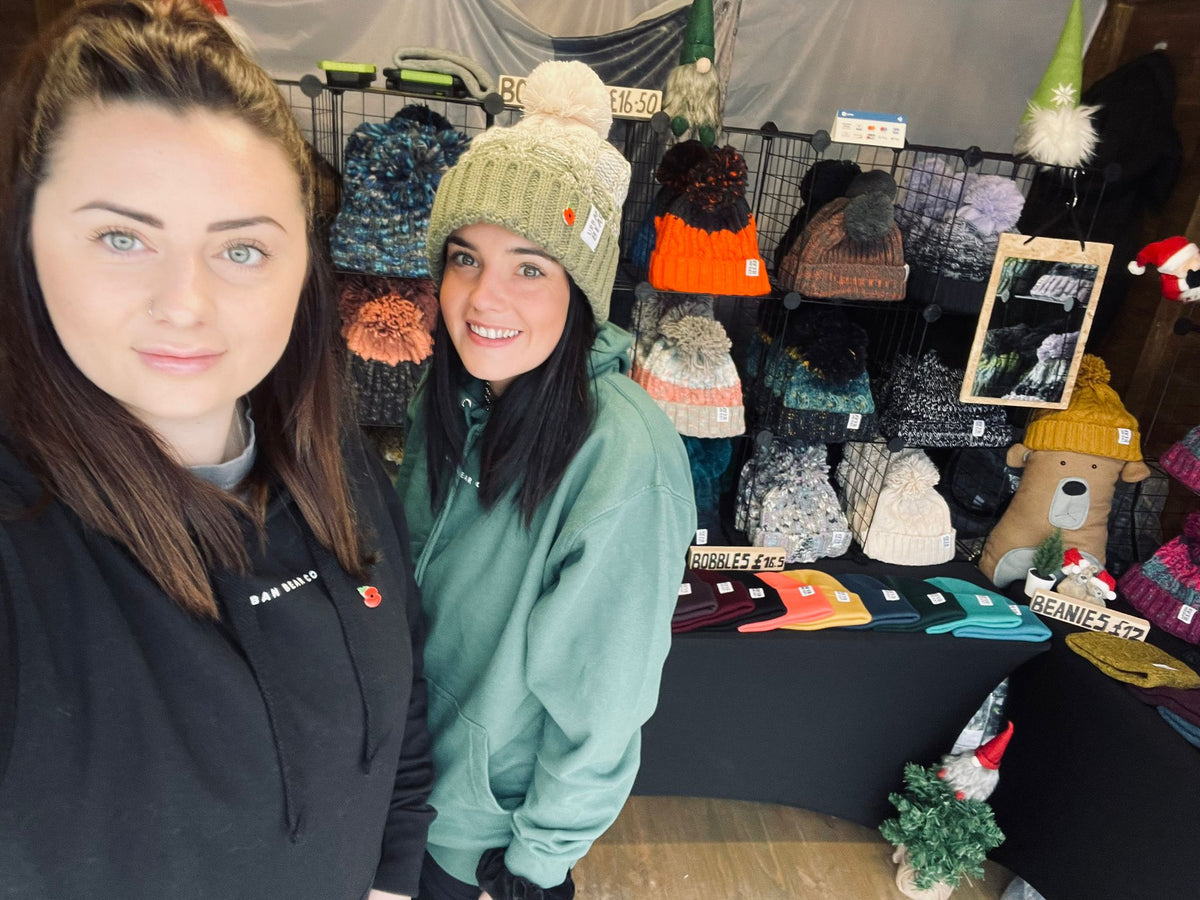 Two women posing at a market stall filled with various knit winter hats, beanies, and small holiday decorations. One woman is wearing a green beanie with a pom-pom, and the other is wearing a black hoodie.