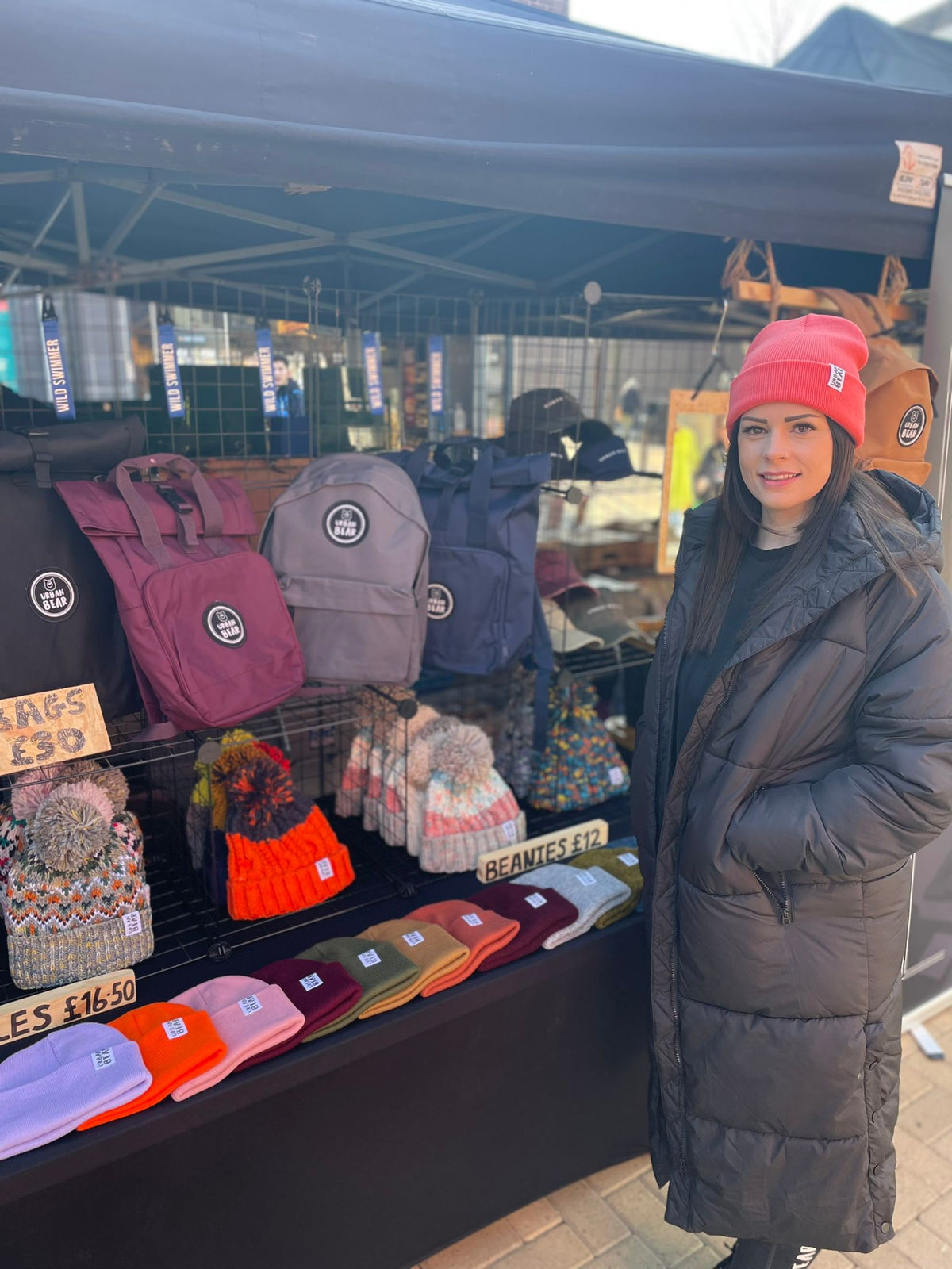 A woman wearing a pink beanie and a long black puffer coat stands next to an outdoor market stall displaying various colorful backpacks, hats, and scarves.