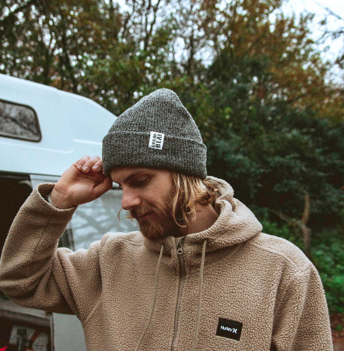 A man wearing a gray knit beanie and a beige sherpa hoodie, both with a visible NAVY K logo patch, standing outdoors in front of a white camper van and trees.