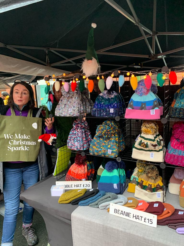 A woman stands at a market stall under a black canopy holding a green tote bag that reads 'We Make Christmas Sparkle.' The stall displays many colorful knit beanies and hats hanging on racks and arranged on tables.