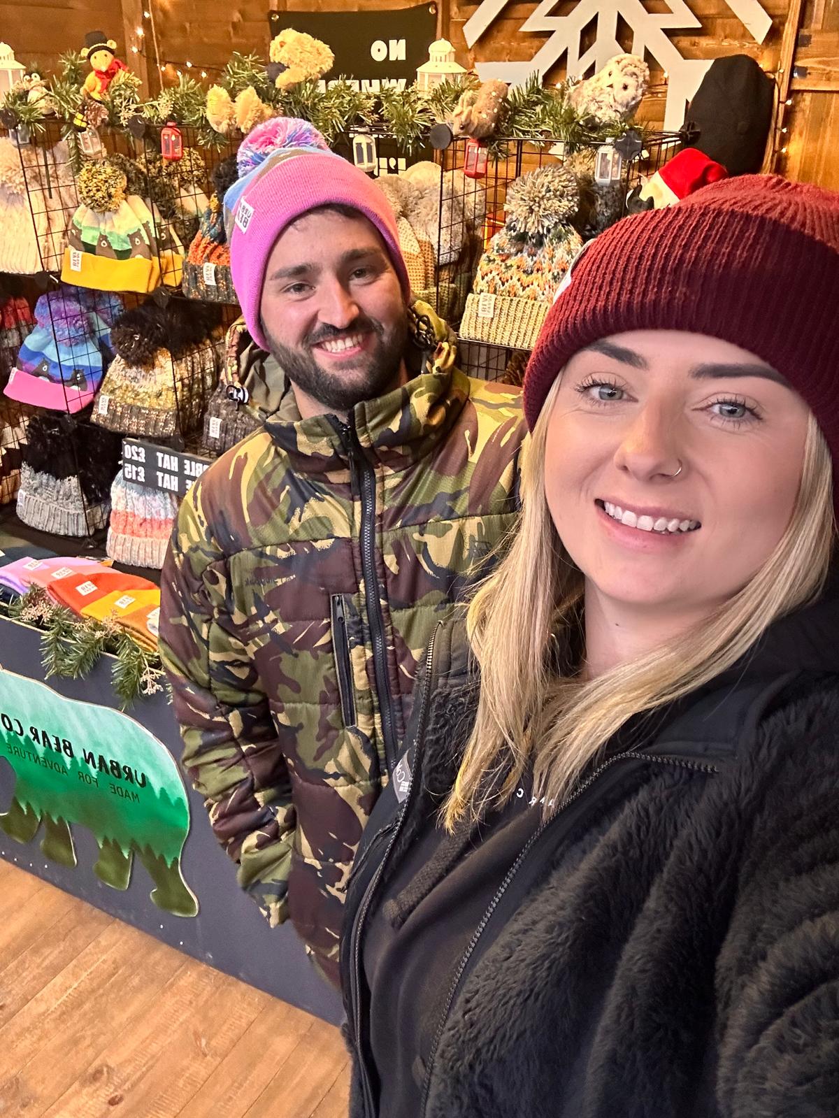 A man and a woman smiling in a cozy, decorated gift shop or market stall. The man wears a pink beanie and a green camouflage jacket, while the woman in the foreground wears a burgundy knit beanie and a black fleece jacket.