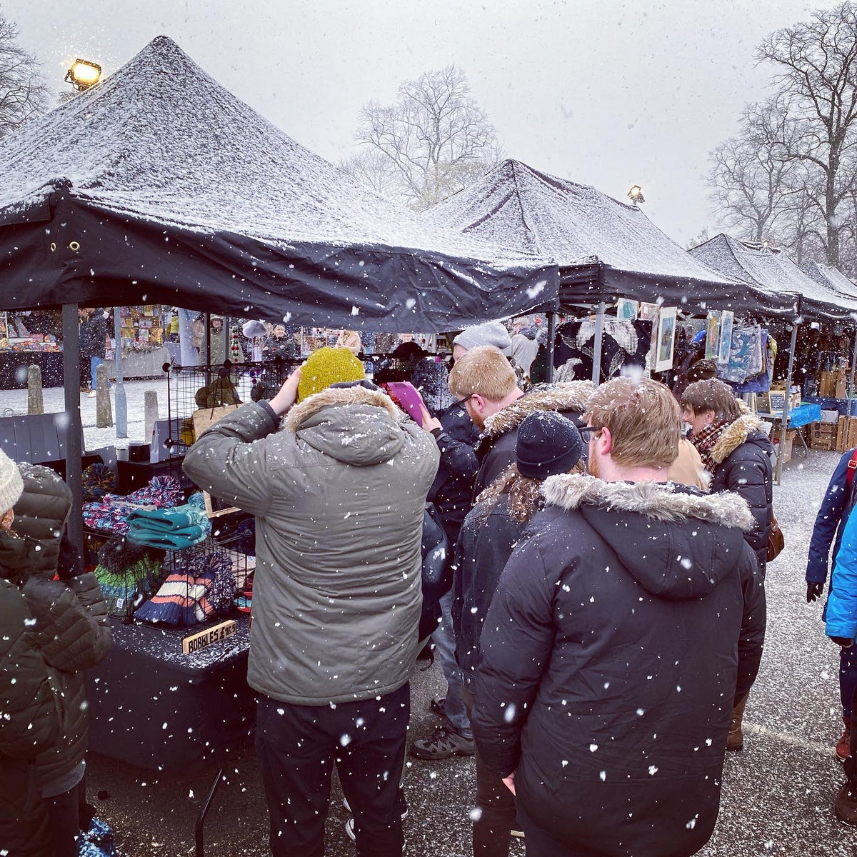 A group of people standing under snow-dusted market tents at an outdoor winter market, with snow falling in the air. Several people are seen from behind, wearing winter jackets and hats, and the scene is set against bare trees in the background.