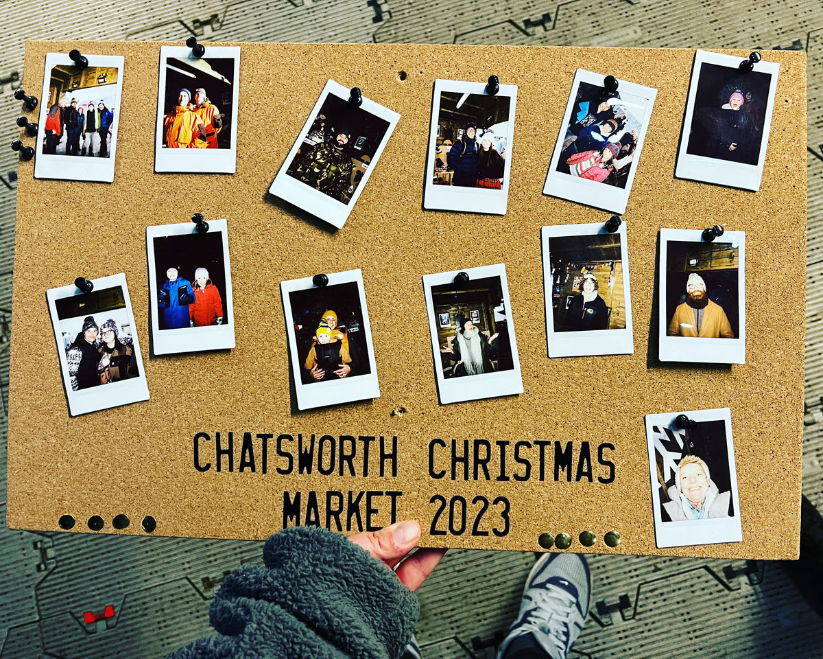 A cork board held by a hand, decorated with multiple Polaroid photos of people at an event and the text 'CHATSWORTH CHRISTMAS MARKET 2023'.