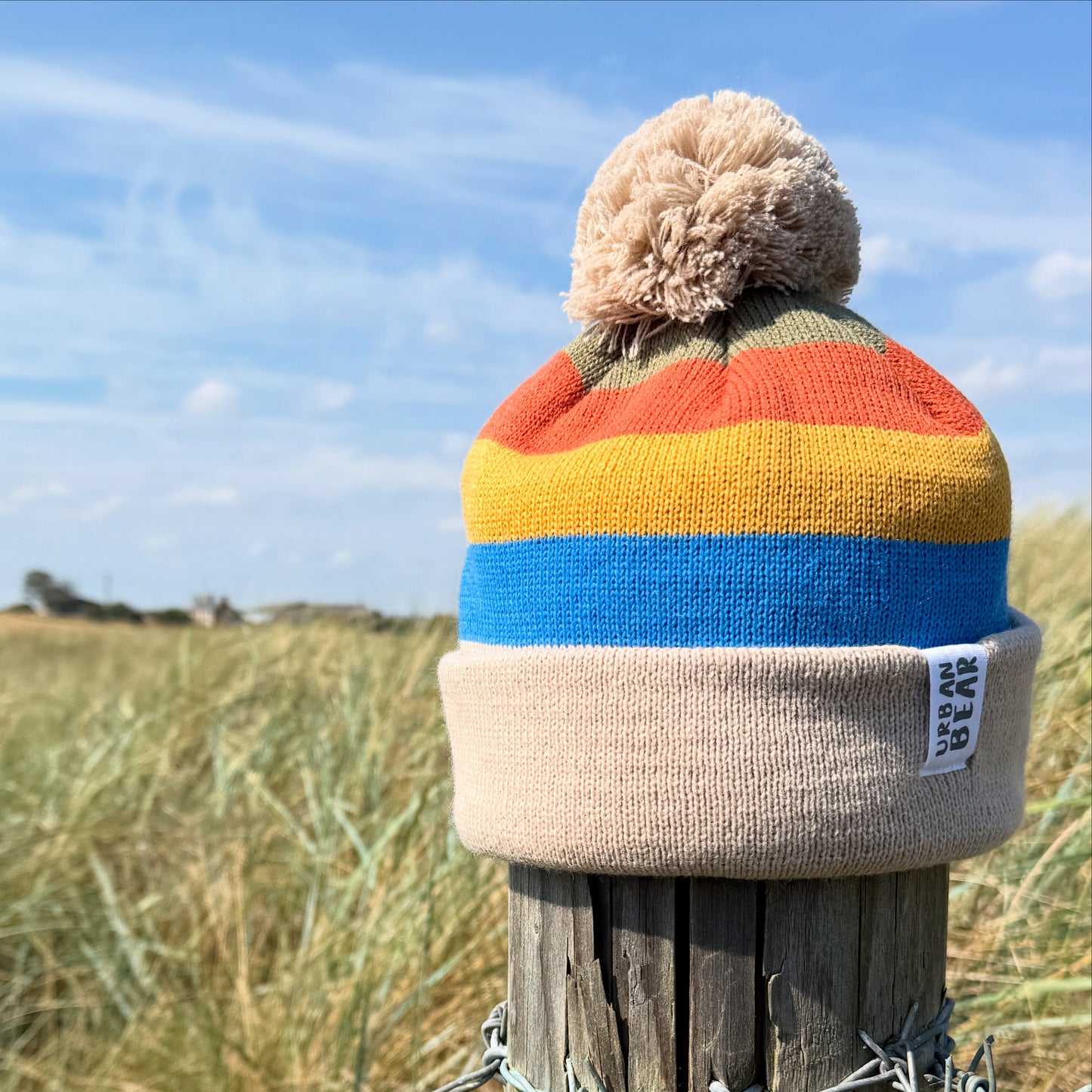 Colorful striped beanie with a pom-pom on a wooden post against a blue sky and grassy field.