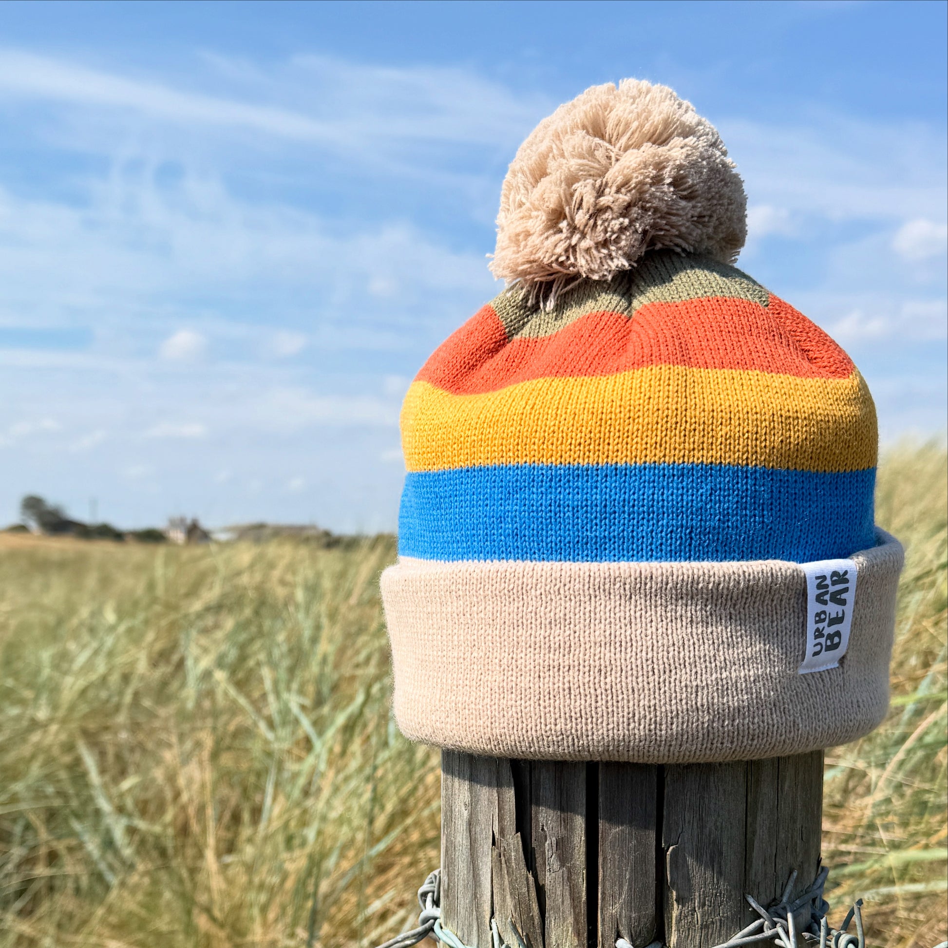 Colorful striped beanie with a pom-pom on a wooden post against a blue sky and grassy field.