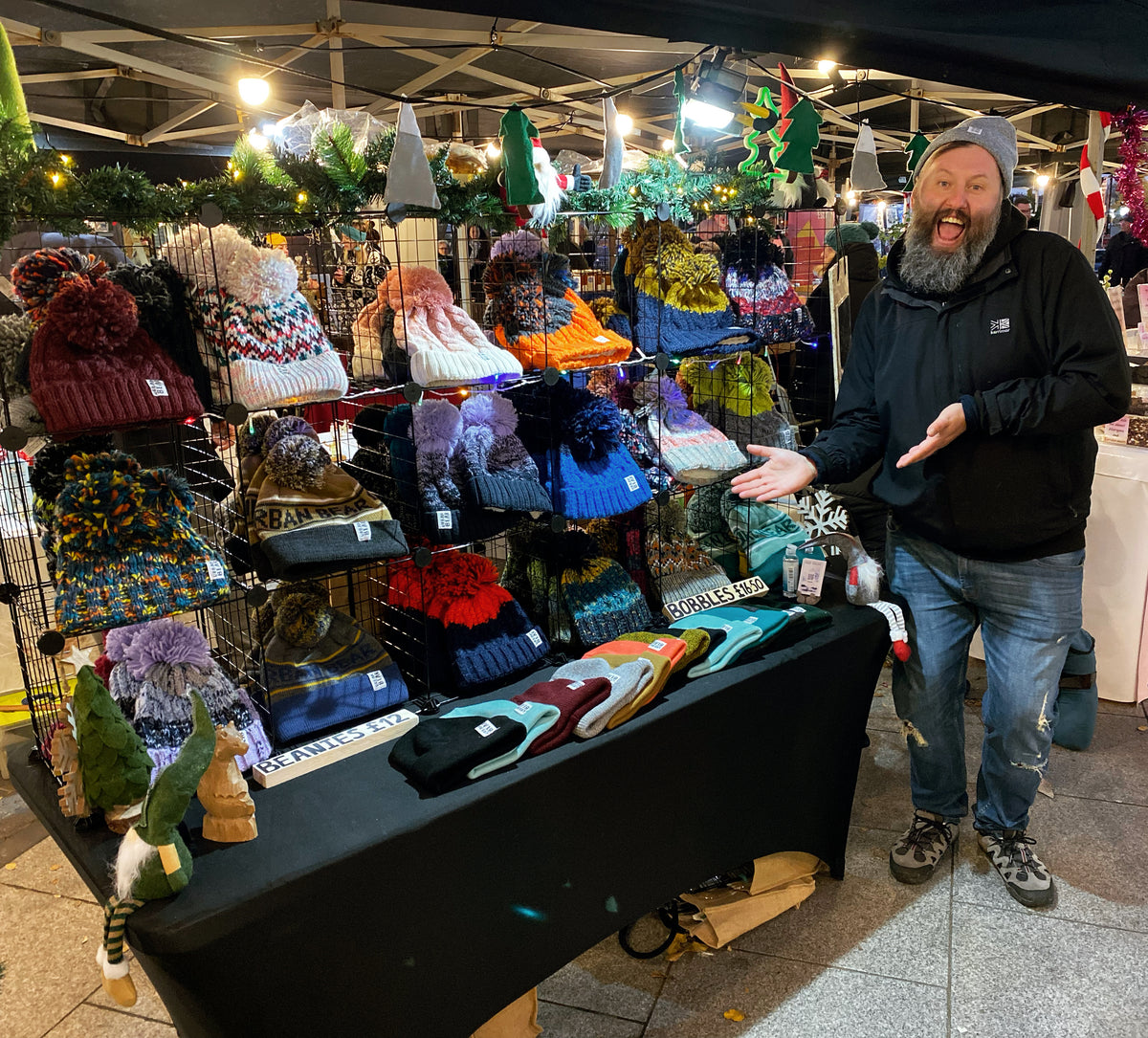 A smiling bearded man in a black jacket and gray beanie stands beside a market stall filled with colorful knit winter hats, scarves, and small green decorative figures under overhead lights.