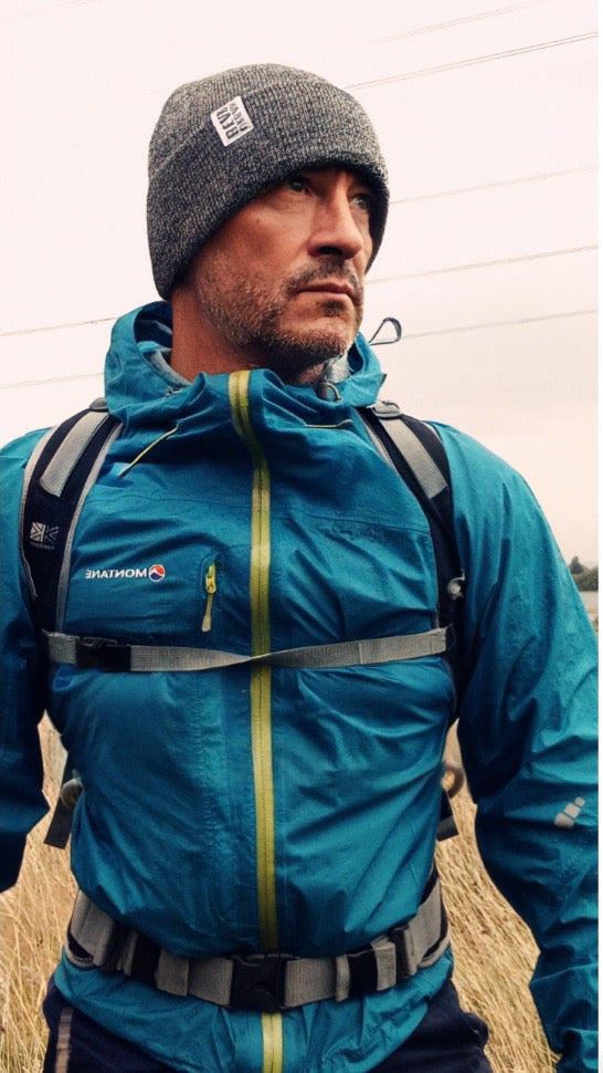 A man wearing a teal Salomon waterproof jacket with yellow zipper accents, a gray knit beanie, and a backpack, standing outdoors in a field.