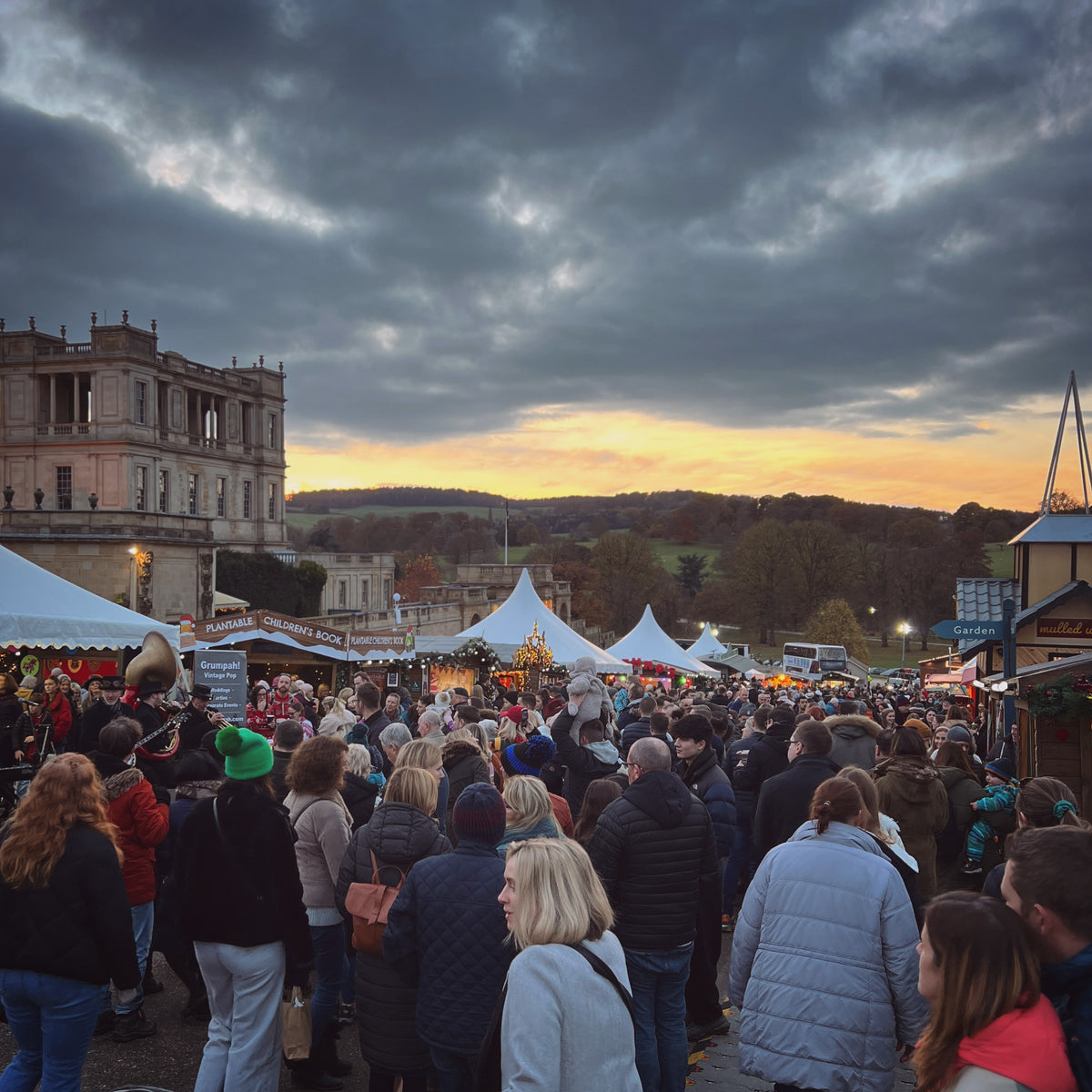 A crowded outdoor market scene at dusk with white tents, stalls, people walking, hills in the distance, and a large historic building under dramatic cloudy skies.