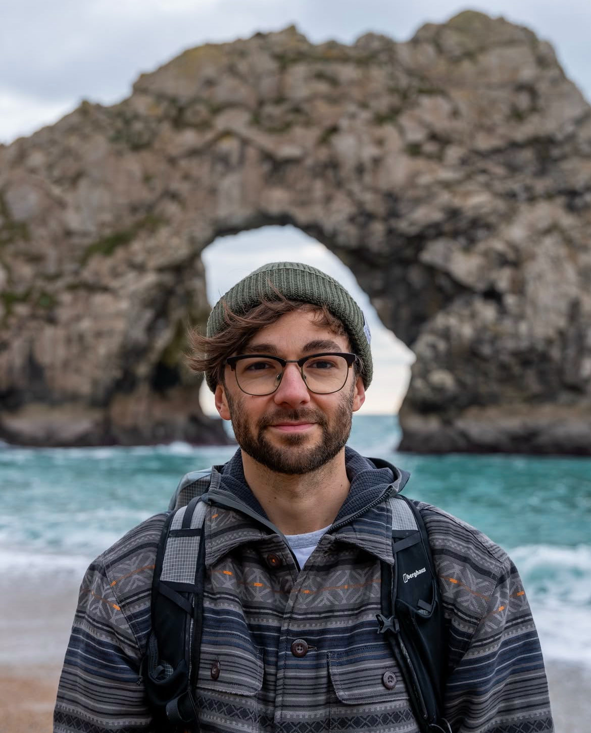 A man wearing a green knit beanie, glasses, and a patterned button-up shirt stands on a beach with a large natural rock arch and turquoise ocean water behind him.