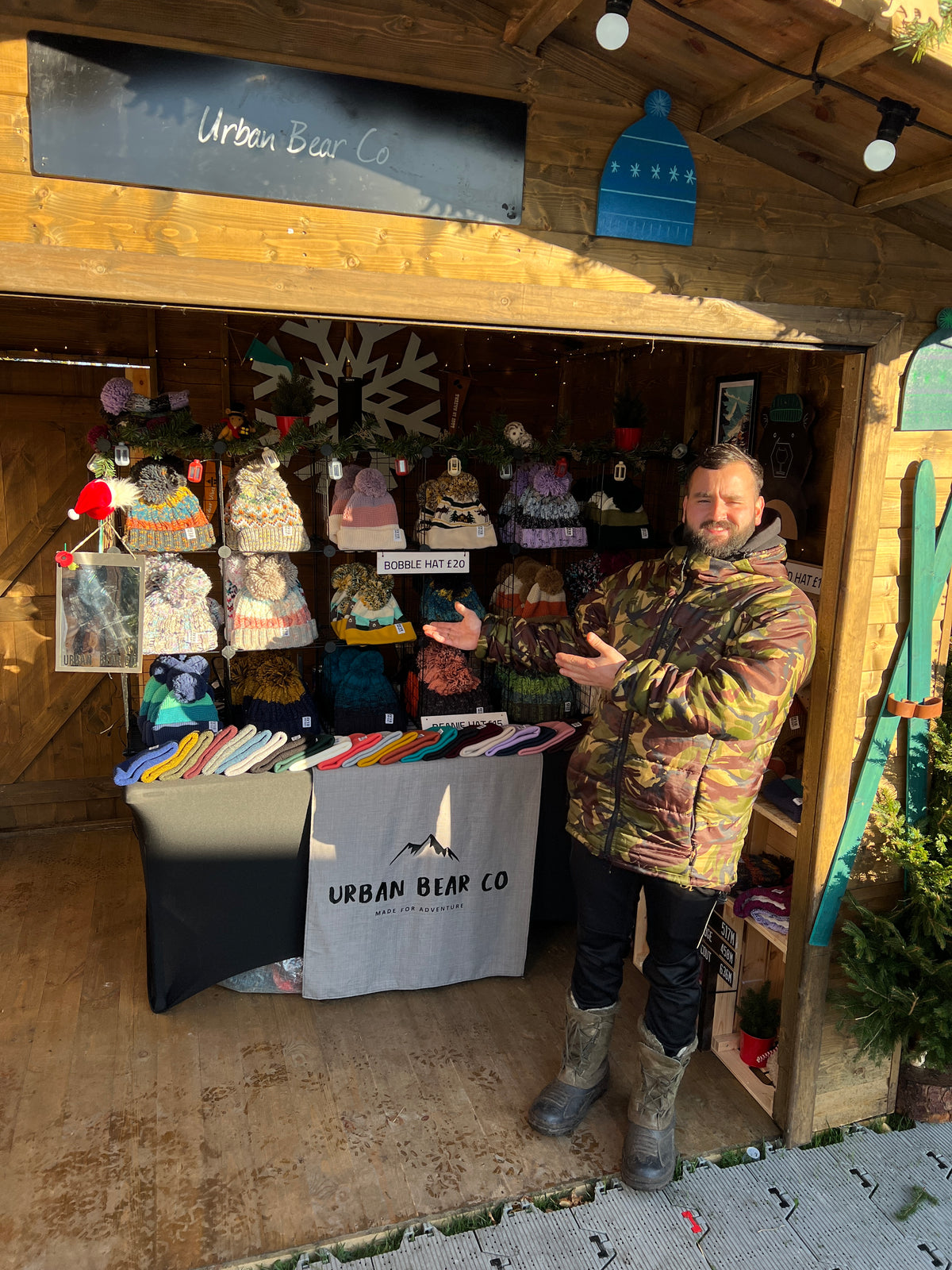 A man wearing a camouflage puffer jacket and gray boots stands in front of a wooden market stall displaying many knit hats and scarves, with a sign reading 'Urban Bear Co' above and on the stall front.