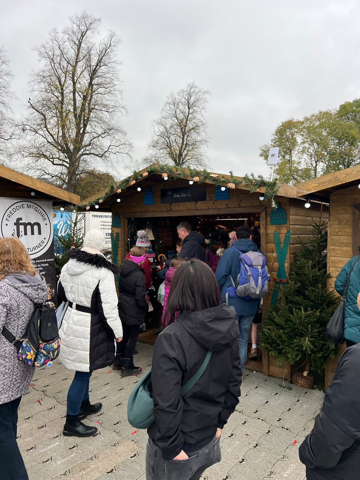 A crowd of people standing outside a small wooden market stall decorated with greenery and string lights, with bare trees and an overcast sky in the background.