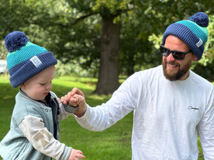 Man and child wearing matching blue beanies with pom-poms in a park setting.
