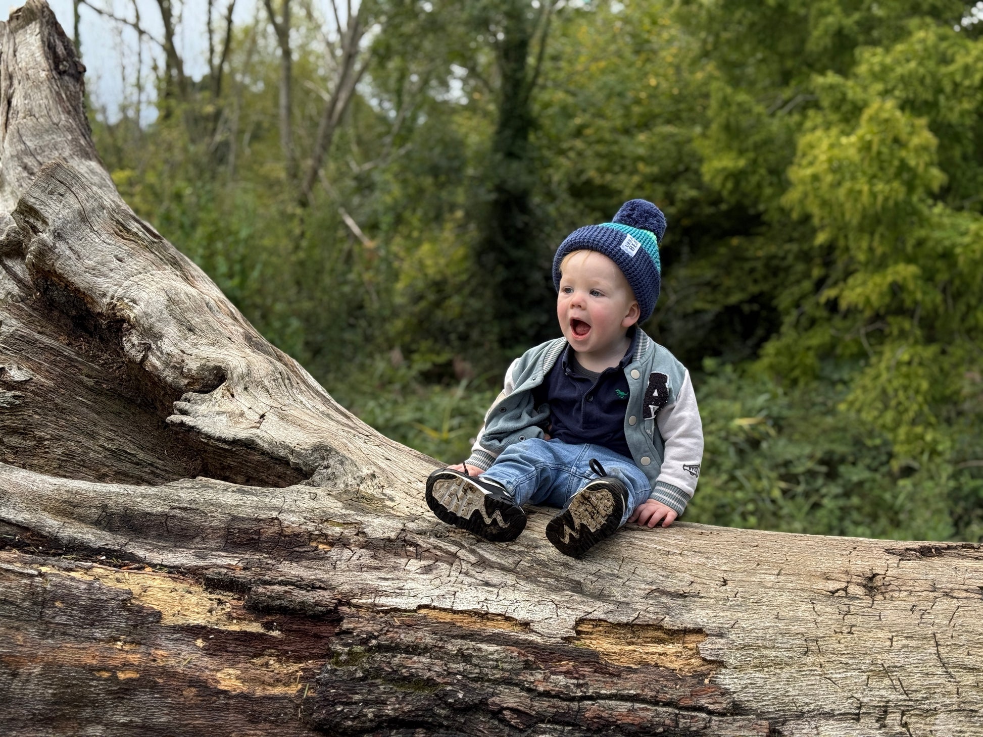 Child sitting on a log in a forest wearing bobble hat