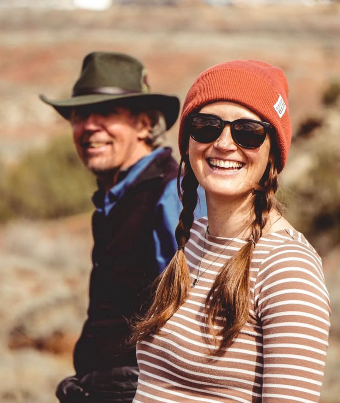 A smiling woman in the foreground wears an orange beanie, sunglasses, and a brown-and-white striped shirt, while a man in the background wears a wide-brimmed hat and blue shirt, set against a blurred outdoor desert landscape.