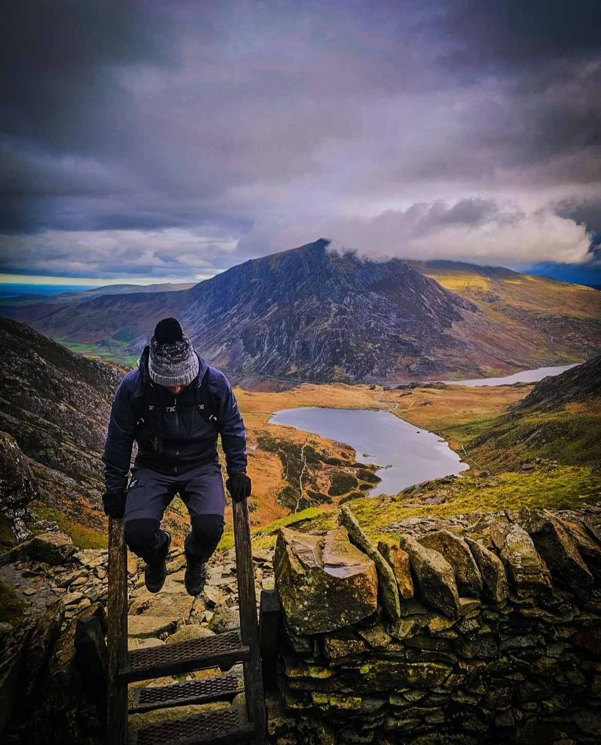 A person in winter clothing climbs a wooden ladder over a stone wall, overlooking a dramatic mountain landscape with a lake under a cloudy sky.