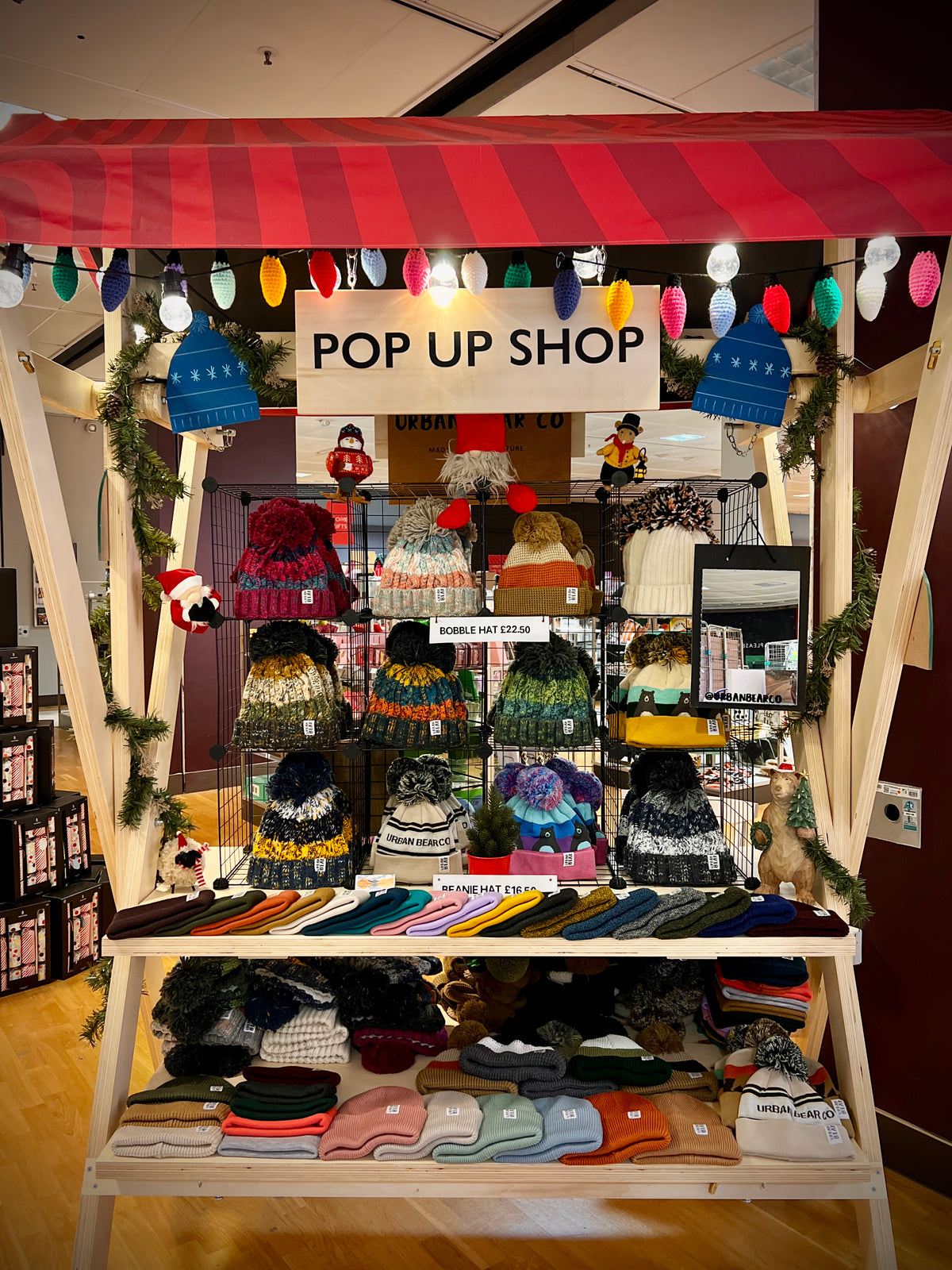 A wooden pop-up display stand covered with a red-and-white striped awning and decorated with holiday lights and greenery, showcasing a wide variety of knit hats, scarves, and folded sweaters under a sign that reads 'POP UP SHOP'.