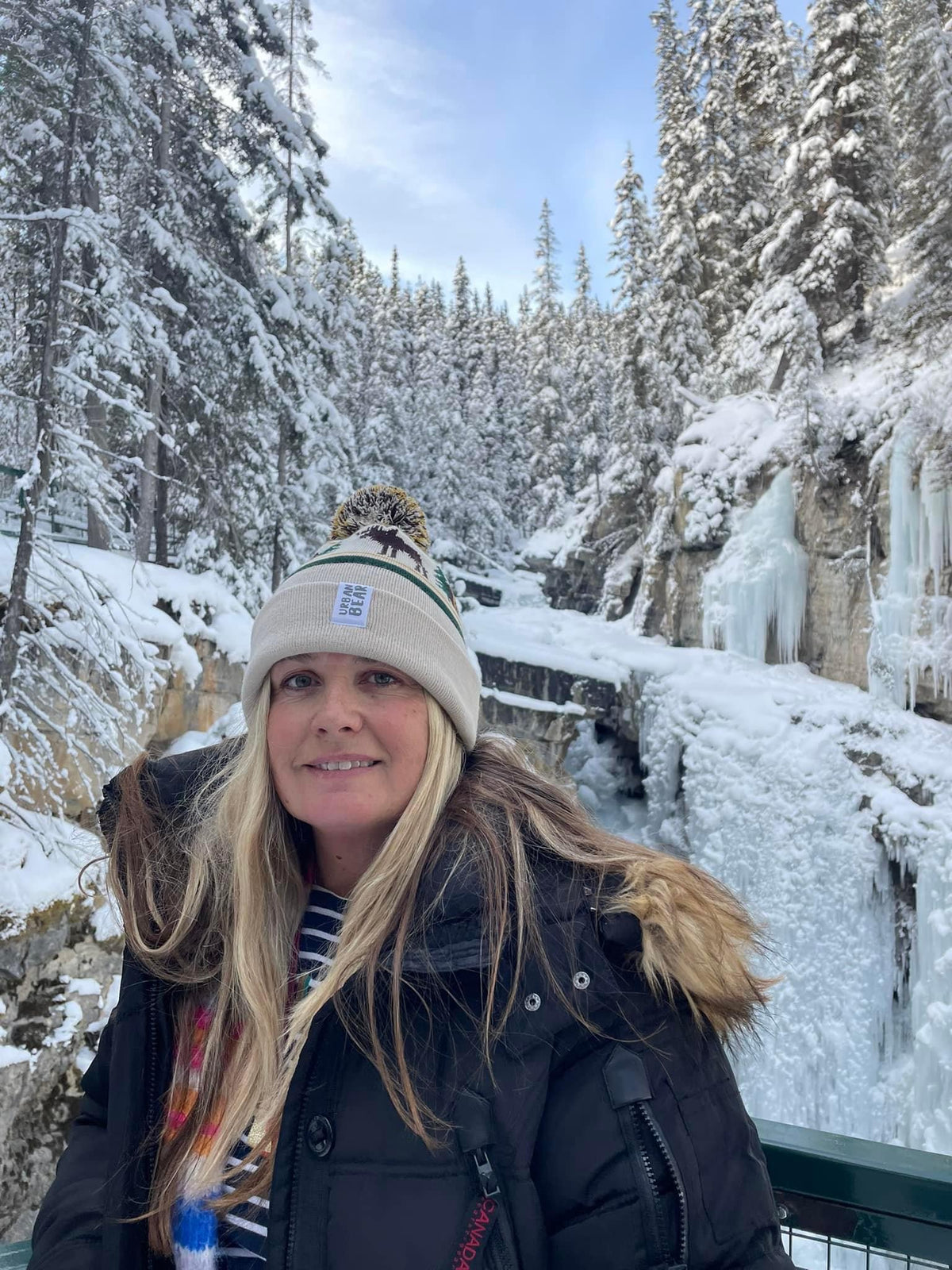 A woman wearing a black winter jacket and a cream knit beanie with a pom-pom stands outdoors in a snowy mountain setting with frozen waterfalls and snow-covered evergreen trees.