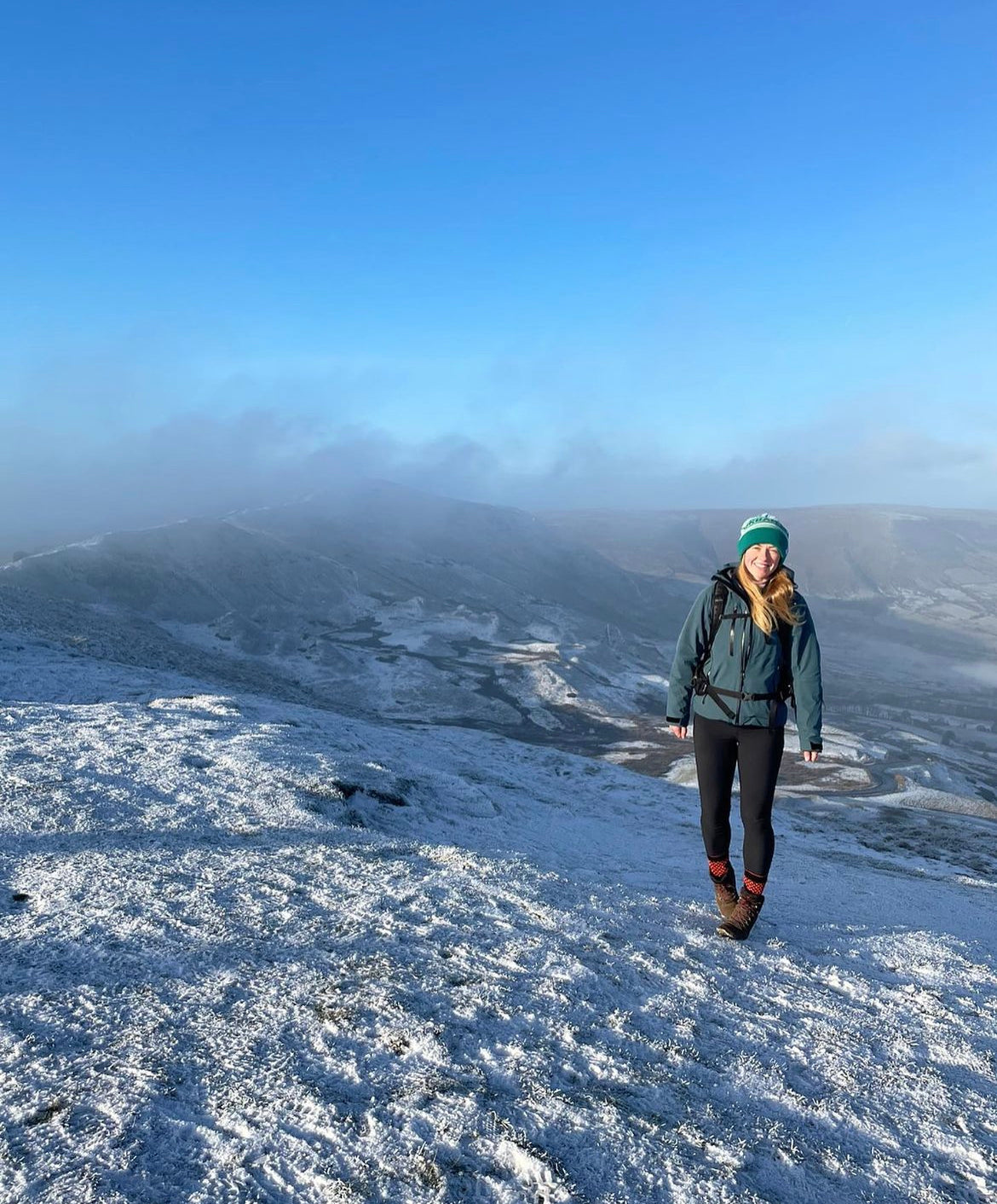 A woman hiking on a snowy mountain ridge under a clear blue sky, wearing a teal jacket, black leggings, and a green beanie.