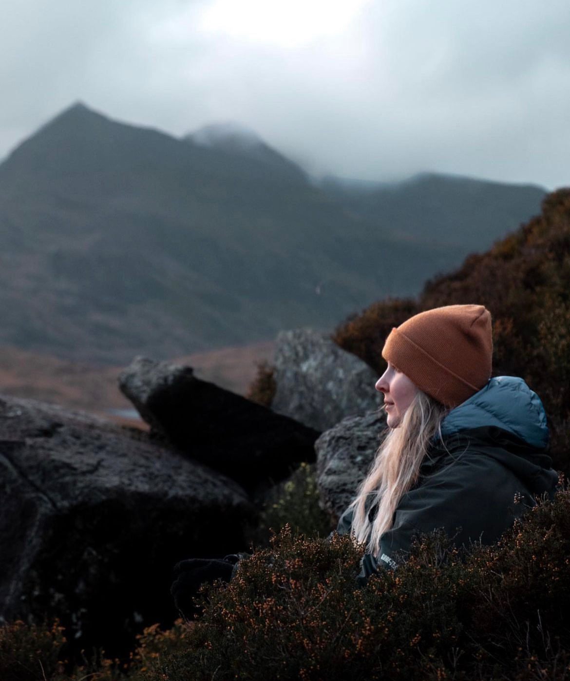 A woman in a tan beanie and dark jacket sits outdoors among rocks and low shrubs, looking toward misty mountains in the background.