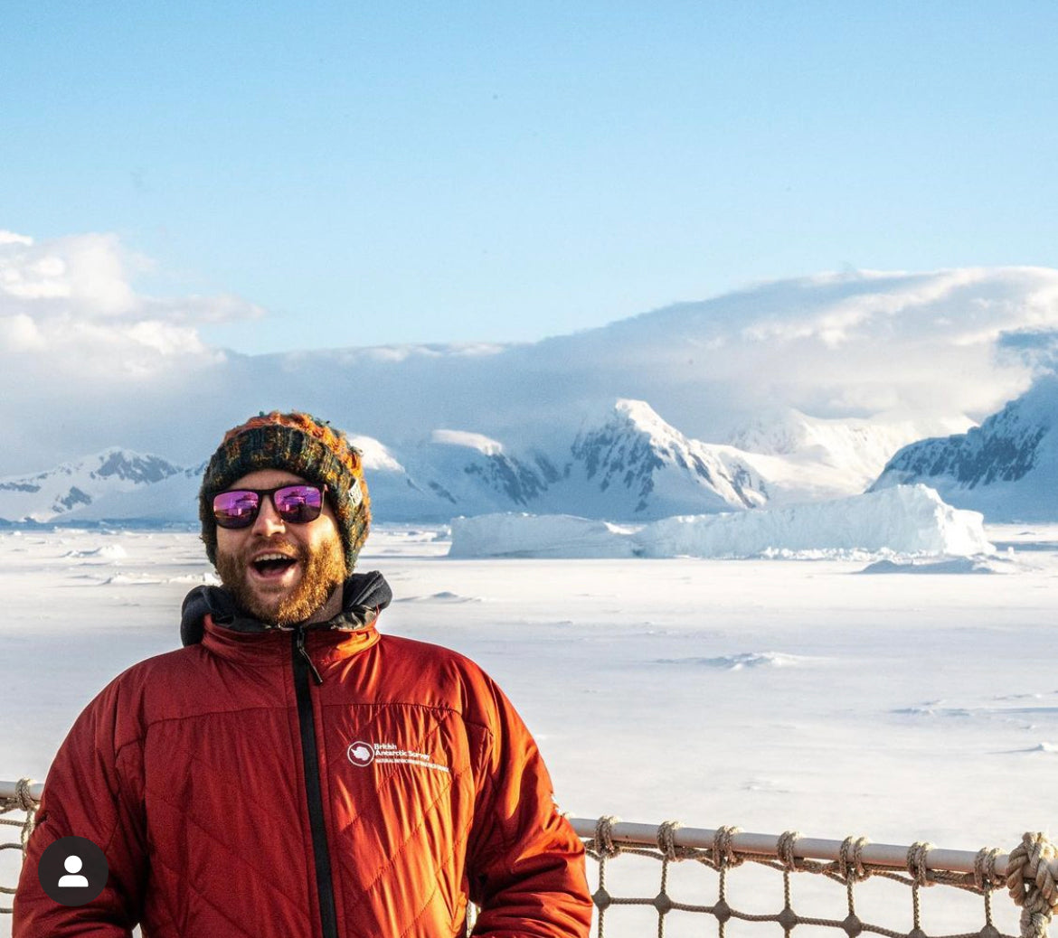 A man wearing a bright orange-red puffer jacket, a knit beanie, and sunglasses stands smiling in front of a snowy mountain landscape under a blue sky.
