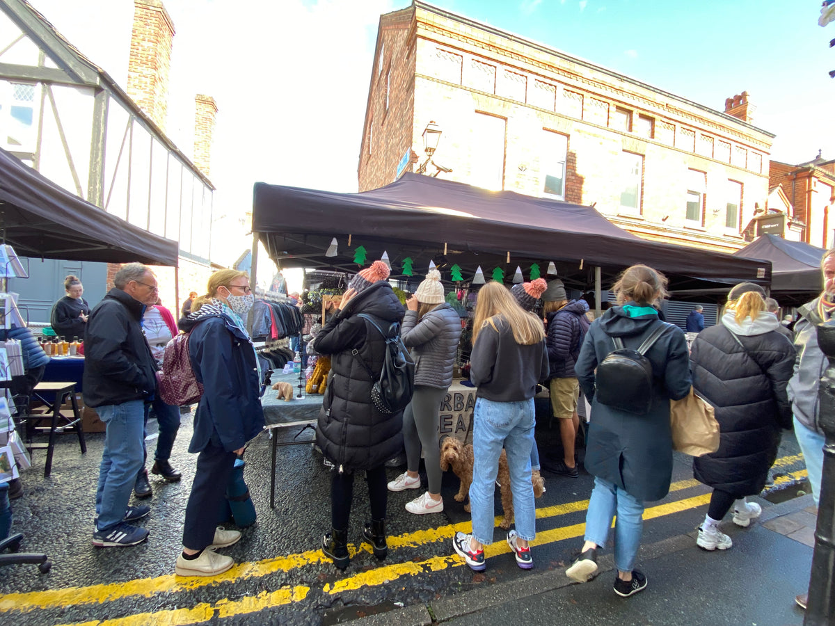A bustling outdoor market scene with multiple people standing under black market tents on a street, with brick buildings in the background.
