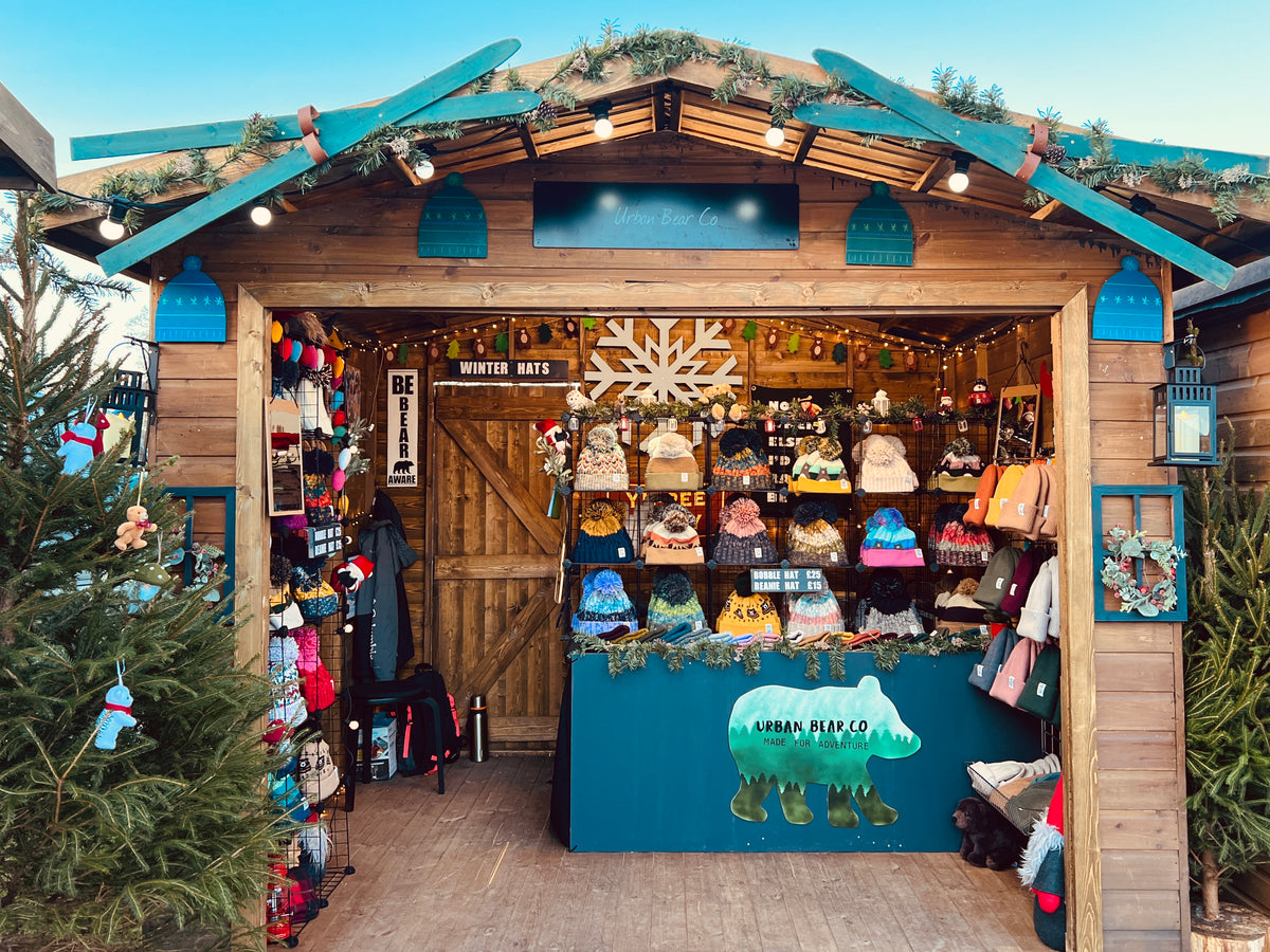 A rustic wooden market stall decorated for winter, with a blue roof, green garlands, and shelves filled with colorful winter hats, scarves, and accessories. A blue counter with a polar bear logo sits in front, and Christmas trees frame the stall against a blue sky.