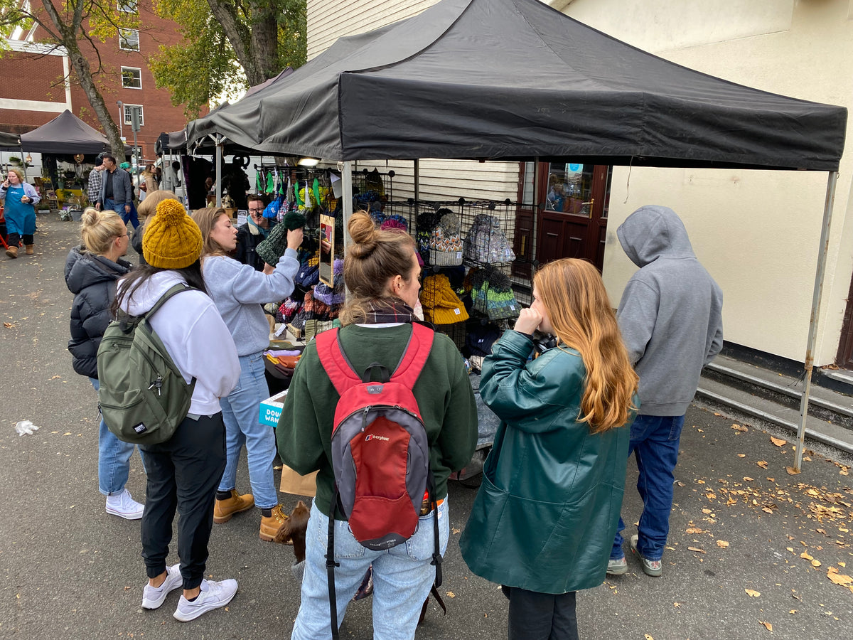 A group of people standing and browsing at an outdoor market stall under a black canopy tent, with clothing and accessories displayed on racks.