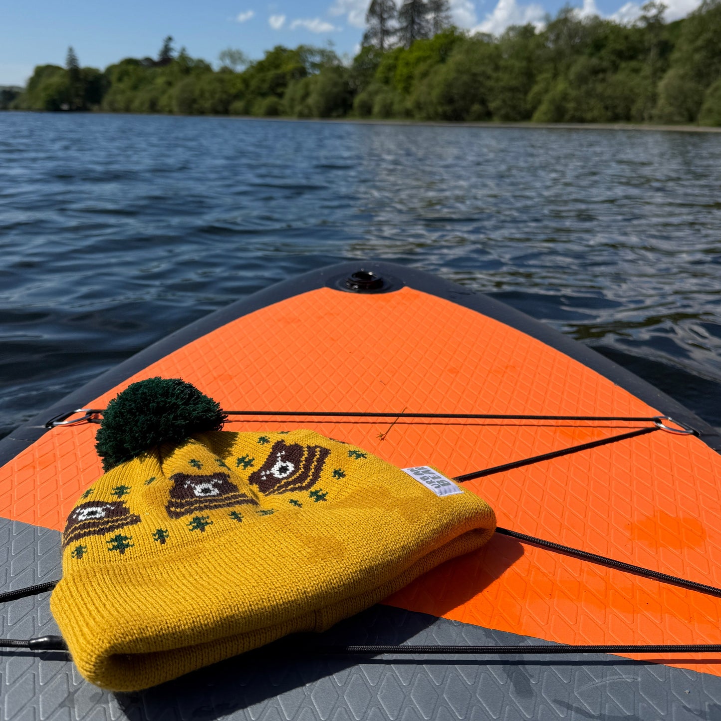 Yellow beanie with bear design and pom-pom on a paddle board against a lake and sky background