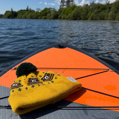 Yellow beanie with bear design and pom-pom on a paddle board against a lake and sky background