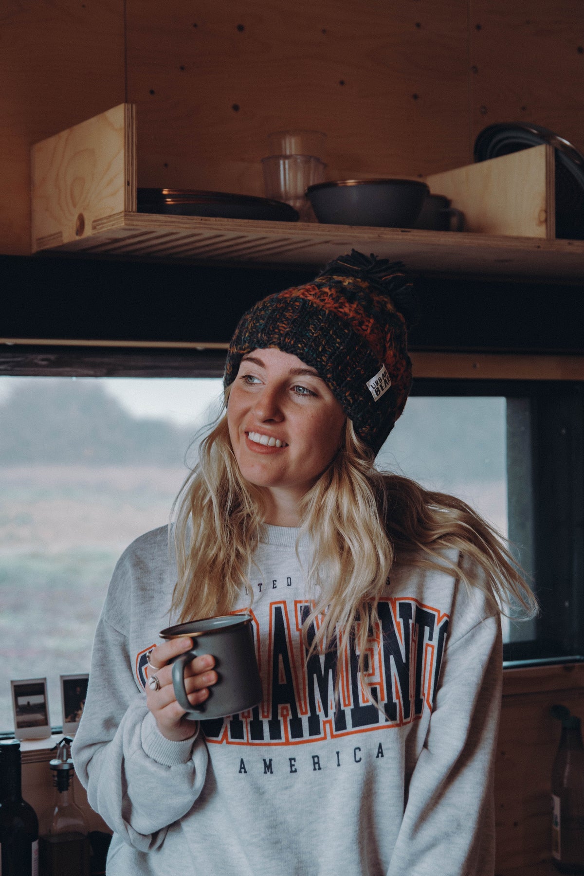 A woman with blonde hair wearing a knit beanie and a light gray sweatshirt with 'The Outfitter America' printed on the front, holding a dark gray mug inside what appears to be a wooden van or camper.