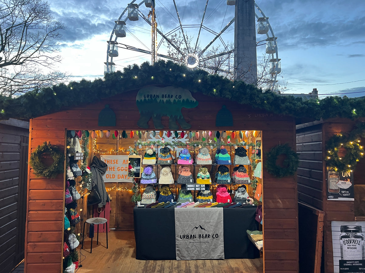 A wooden market stall at an outdoor Christmas market, decorated with green garlands and displaying a variety of colorful winter clothing and accessories. The stall is branded with 'Urban Bear Co' on the sign above the entrance and the tablecloth.