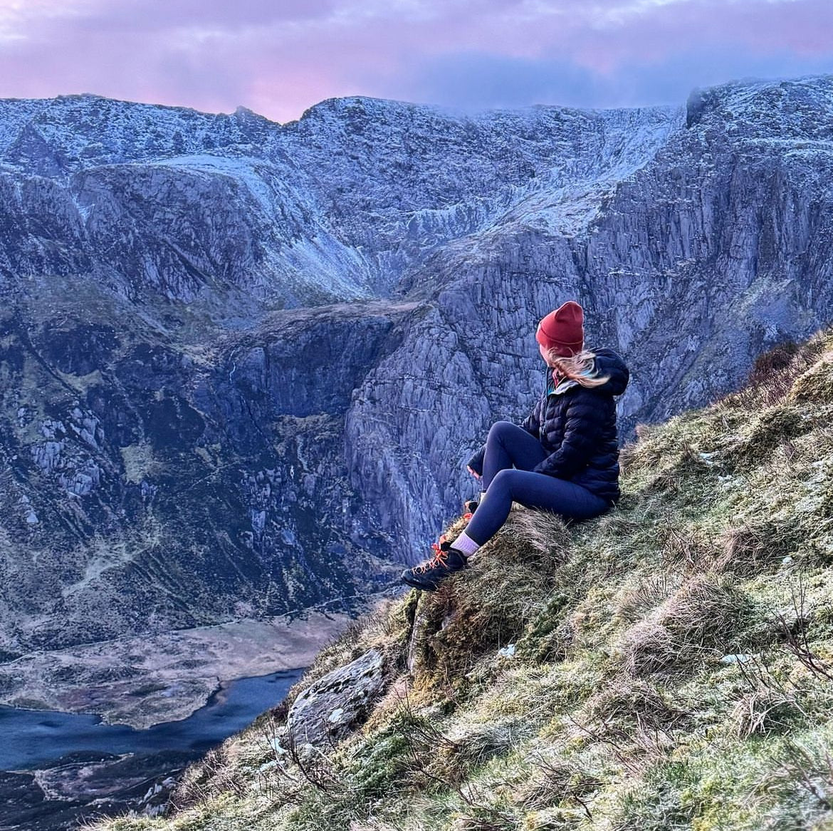 A person wearing a red beanie, black puffer jacket, and dark leggings sits on a grassy mountainside overlooking a deep valley and snowy peaks at dusk.