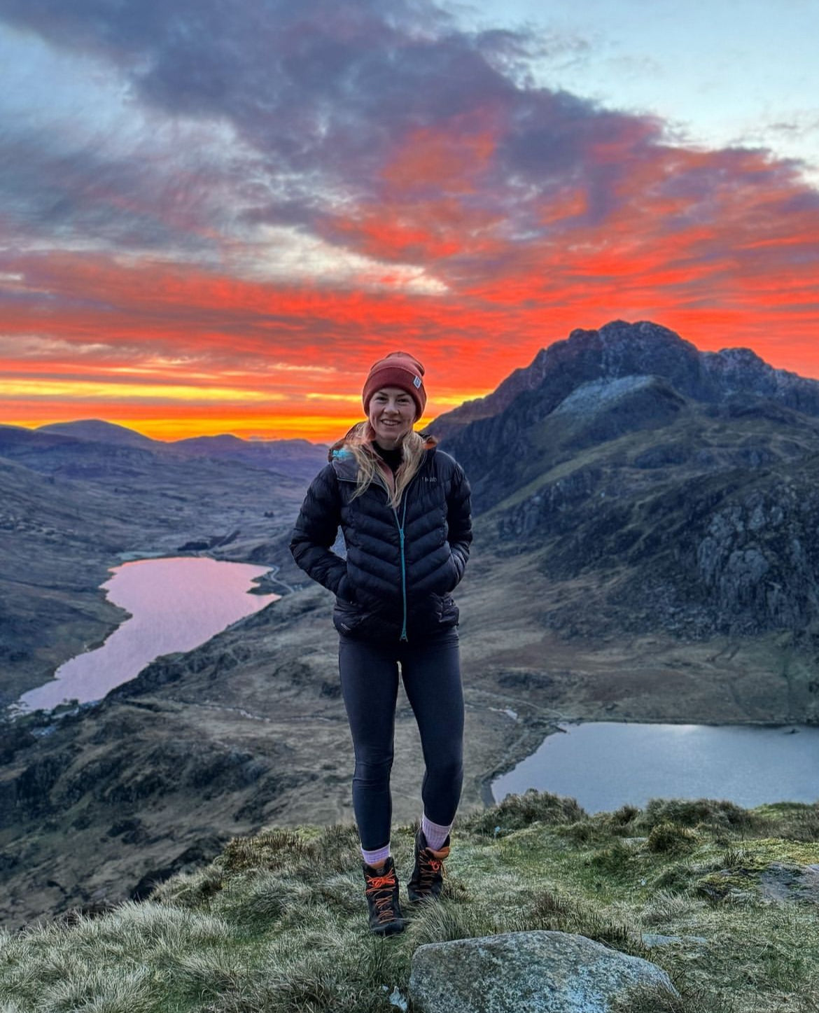 A woman stands on a grassy mountain ridge at sunset, wearing a black puffer jacket, black leggings, and a red beanie, with dramatic orange and pink clouds and lakes in the background.