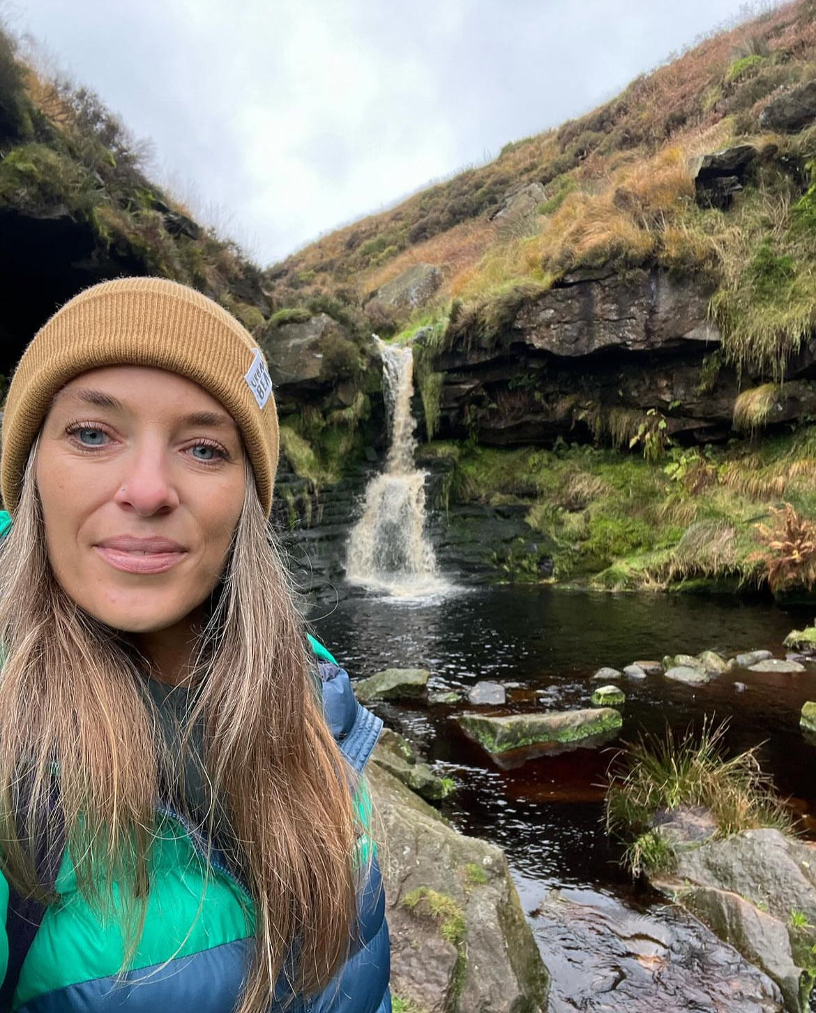 A woman wearing a tan beanie and a green and blue puffer jacket takes a selfie in front of a small waterfall in a rocky, grassy outdoor landscape.