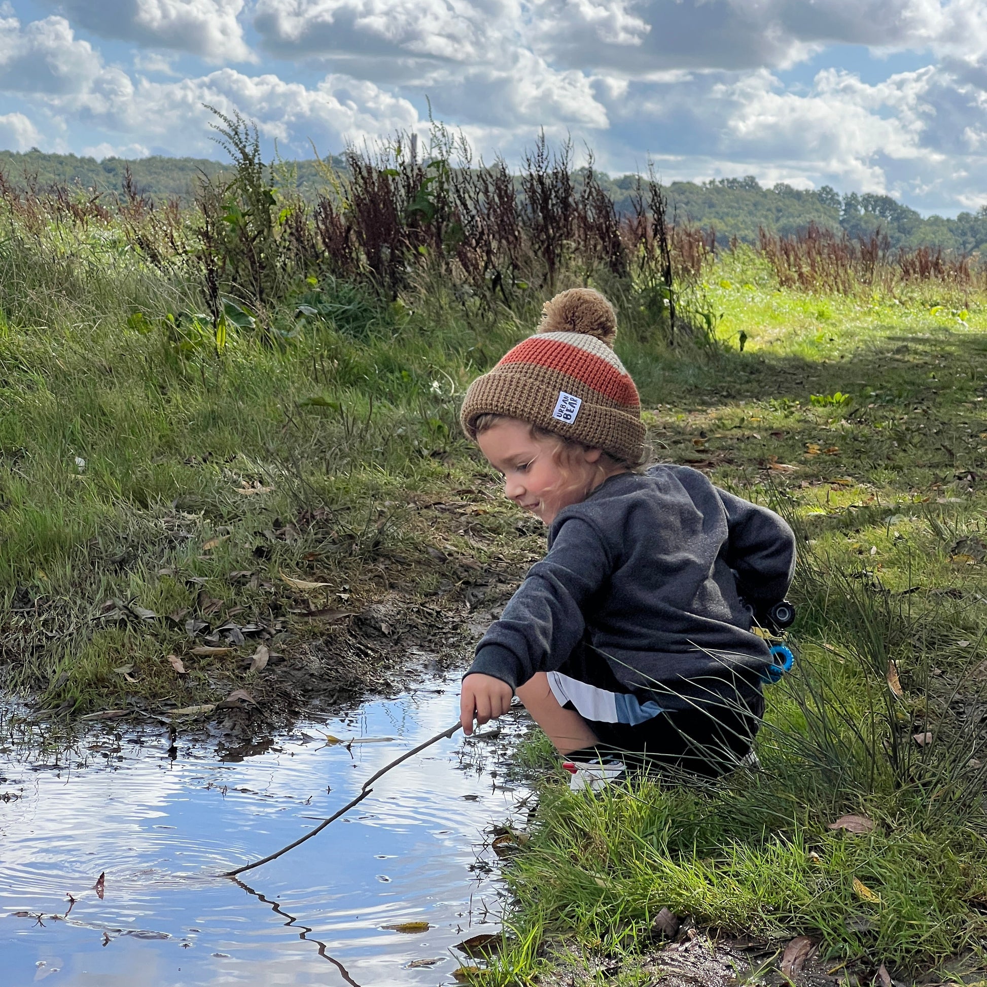 Boy wearing bobble hat holding a stick in a grassy area with a blue sky and clouds.