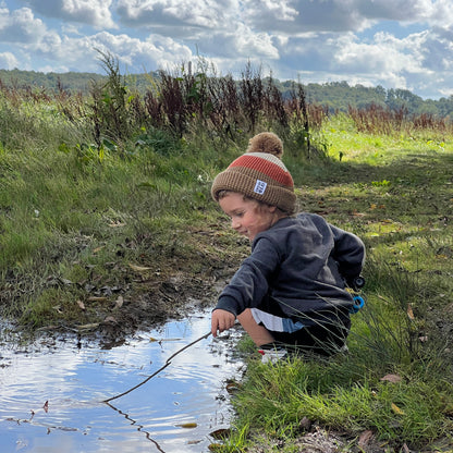 Boy wearing bobble hat holding a stick in a grassy area with a blue sky and clouds.