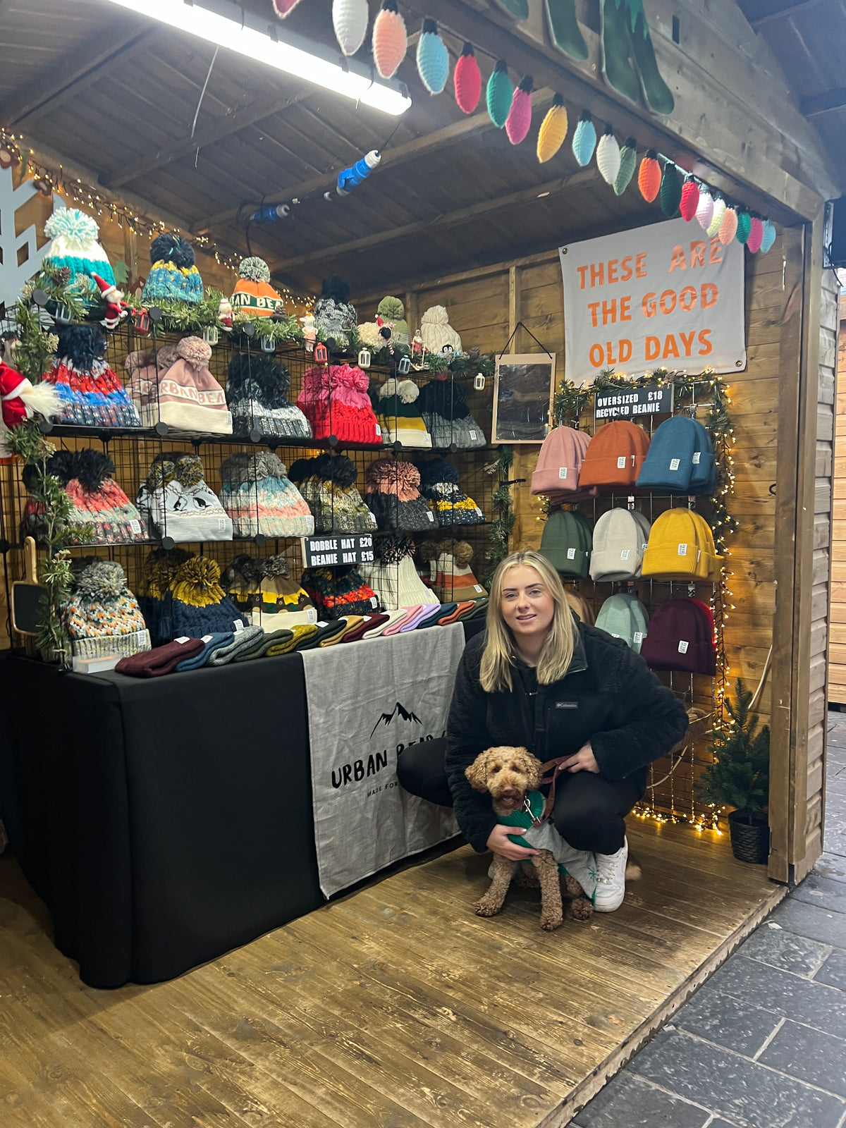 A woman crouches inside a rustic wooden market stall with shelves of colorful knit hats, scarves, and gloves, holding a small tan dog. A sign on the stall reads 'THESE FEEL THE GOOD OLD DAYS'.
