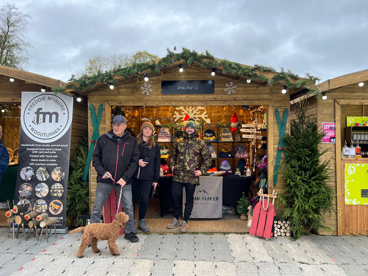 Three people standing in front of a wooden market stall decorated for Christmas, with a small brown dog on a leash in the foreground. A large vertical sign on the left reads 'Falcon Woodworks fm Woodturning'.