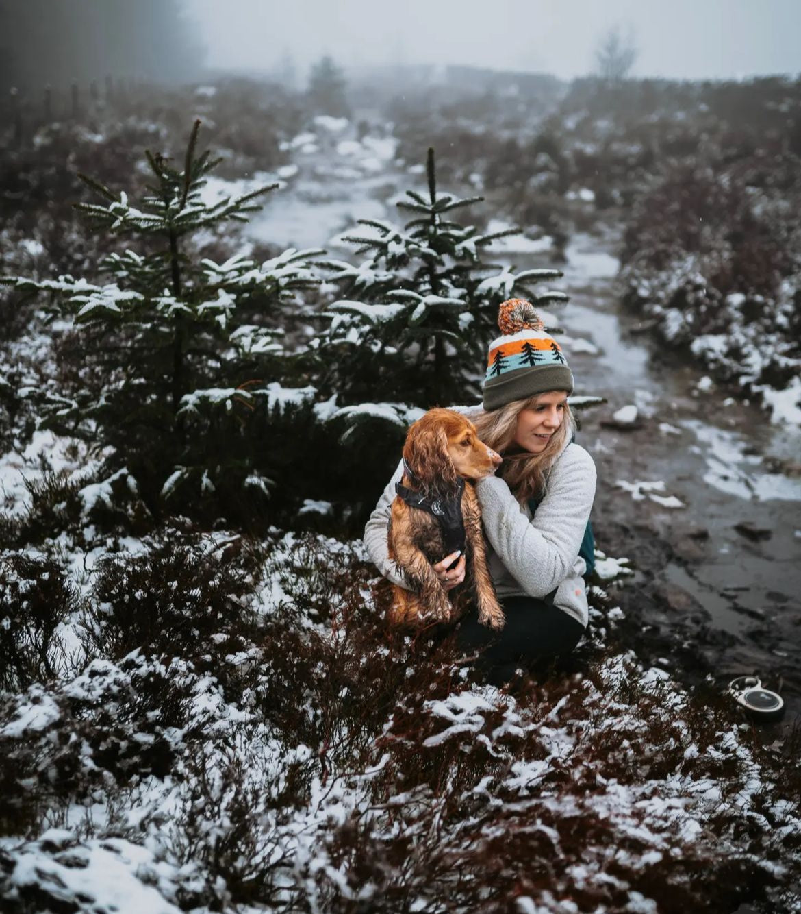 A woman crouches in a snowy outdoor landscape holding a brown dog. She wears a white top, dark pants, and a knit beanie with orange, green, and gray stripes and a pom-pom.