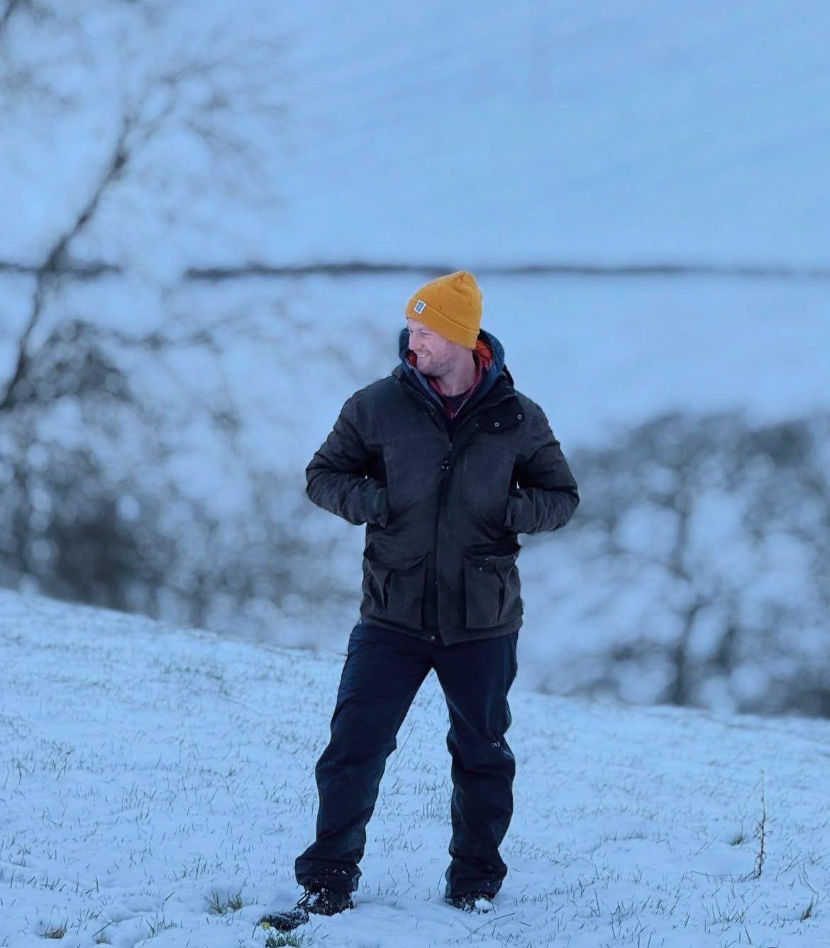 A man standing in a snowy outdoor landscape wearing a dark winter jacket, dark pants, and a mustard yellow knit beanie.