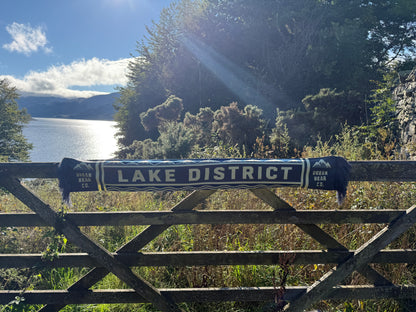 A dark blue and white 'Lake District' scarf draped over a wooden gate, with a scenic view of a lake and forest in the background.