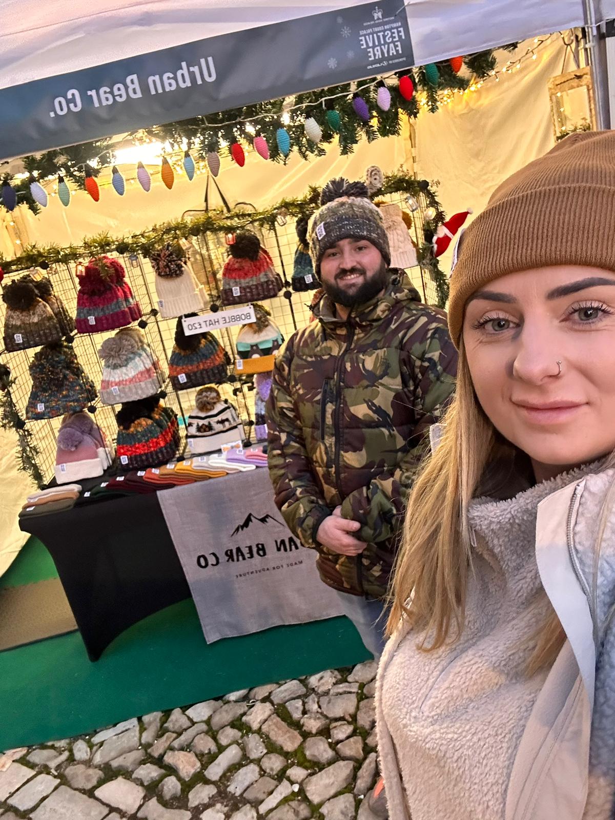 A woman in a brown beanie takes a selfie next to a man in a camouflage winter jacket at an outdoor market booth for Urban Bear Co., with knit hats displayed under a canopy.